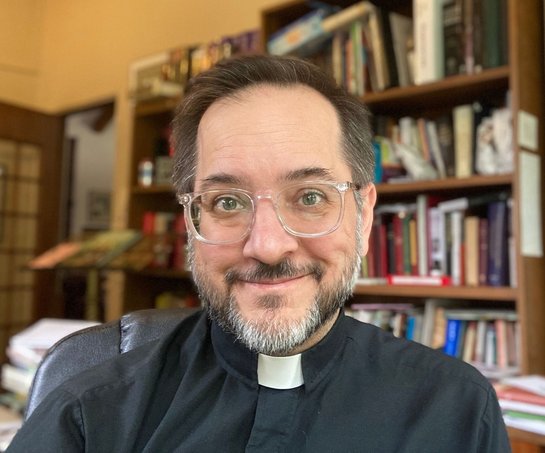 A man with glasses and a beard is sitting in front of a bookshelf.