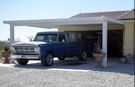 A blue truck is parked under a carport in front of a house.