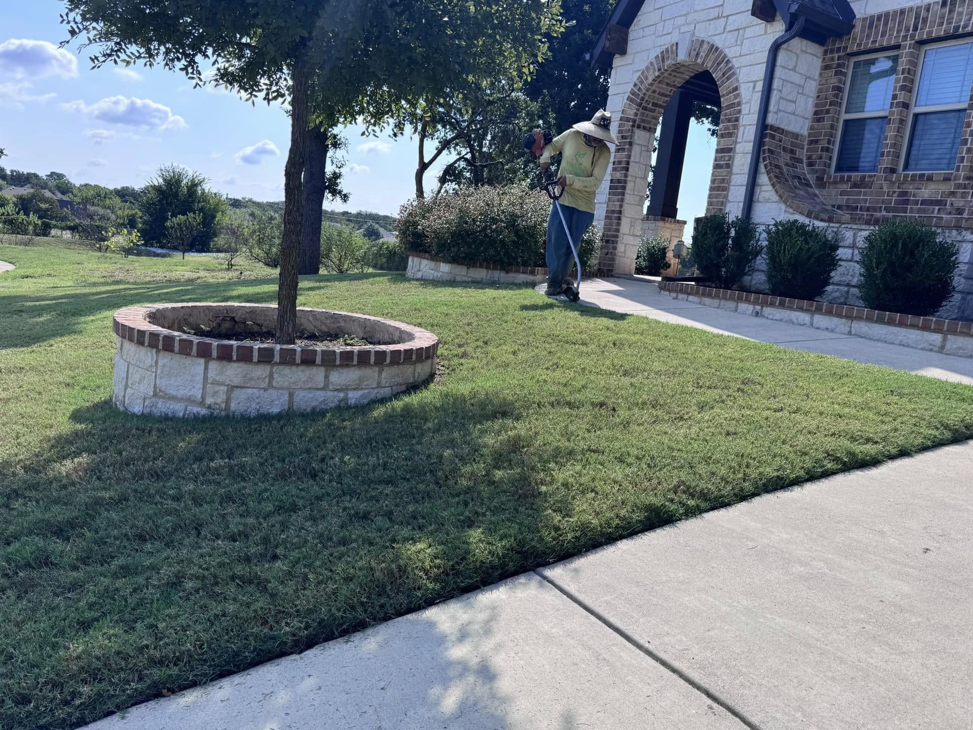 A person mowing a well-manicured lawn near a stone house and tree with a brick planter on a sunny day.