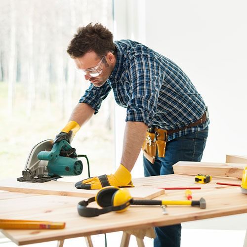 Una persona con gafas y guantes de seguridad utiliza una sierra circular sobre una tabla de madera.