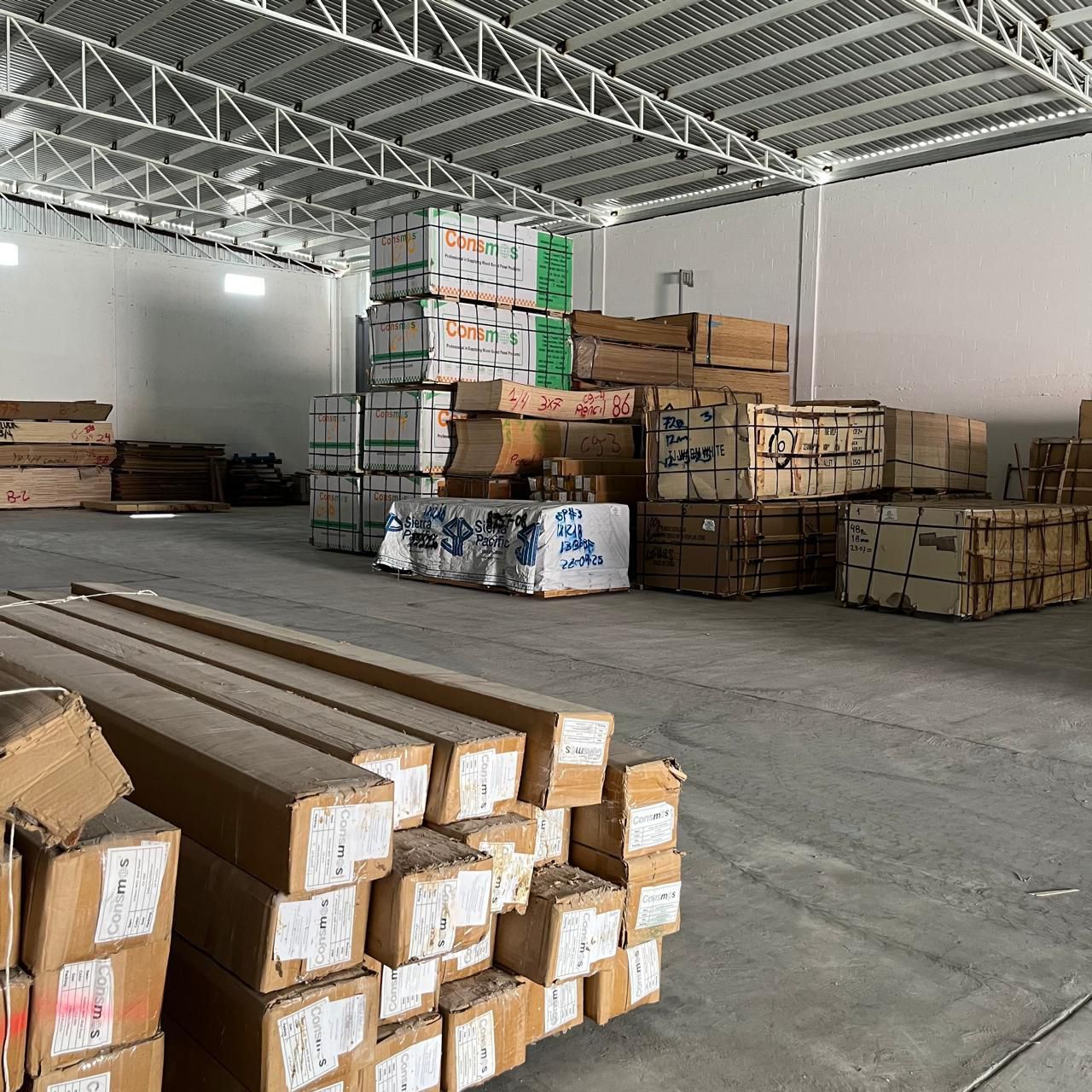 Interior de almacén con pilas de madera y materiales embalados. Paredes y techo blancos, suelo de hormigón.