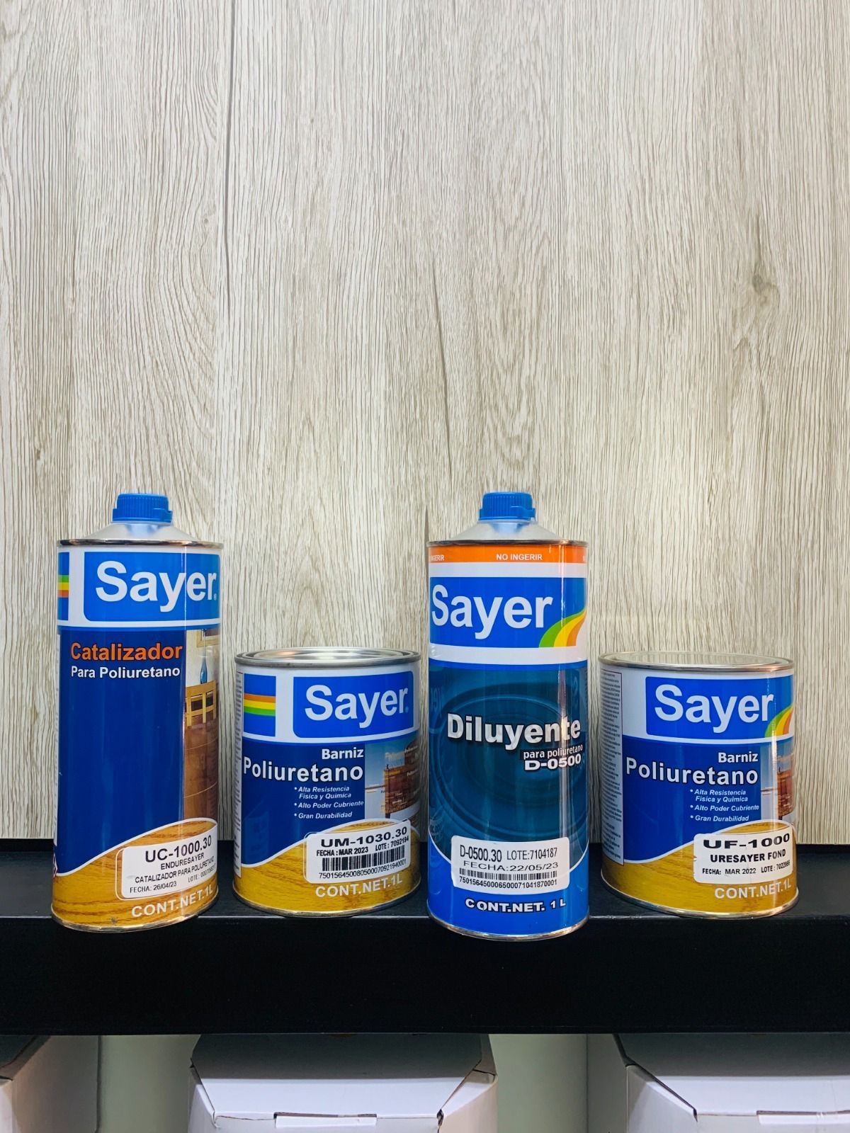 Cuatro latas de productos para el acabado de madera Sayer en un estante, contra un fondo de veta de madera.