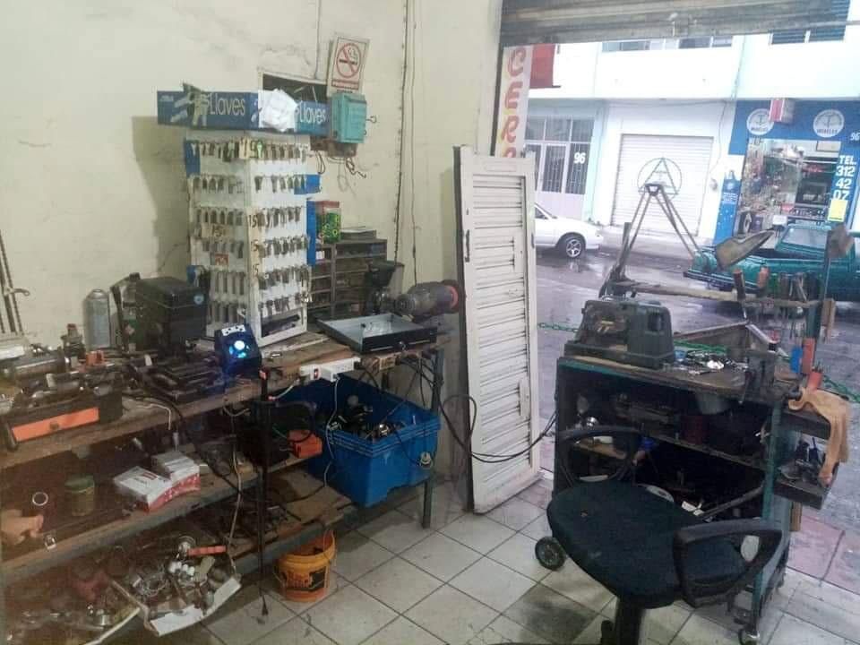 Un interior de cerrajería desordenado con herramientas, estantes para llaves y una puerta abierta a la calle.