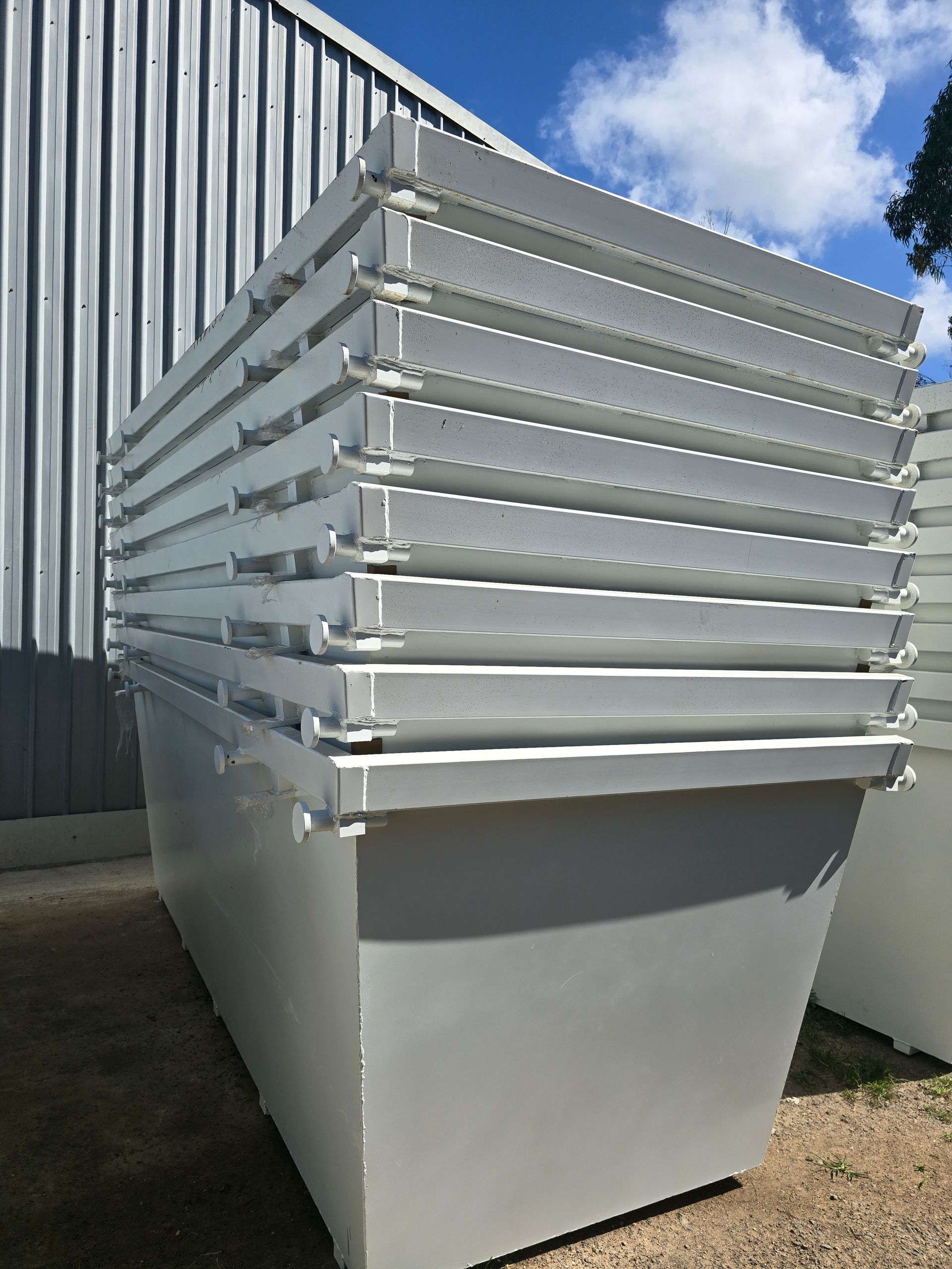 Stack of white metal rectangular bins against a building — Coastal Skip Bin Hire In Craignish, QLD