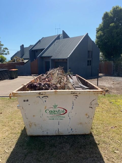 White dumpster overflowing with debris in front of a gray house on a sunny day. — Coastal Skip Bin Hire In Eli Waters, QLD