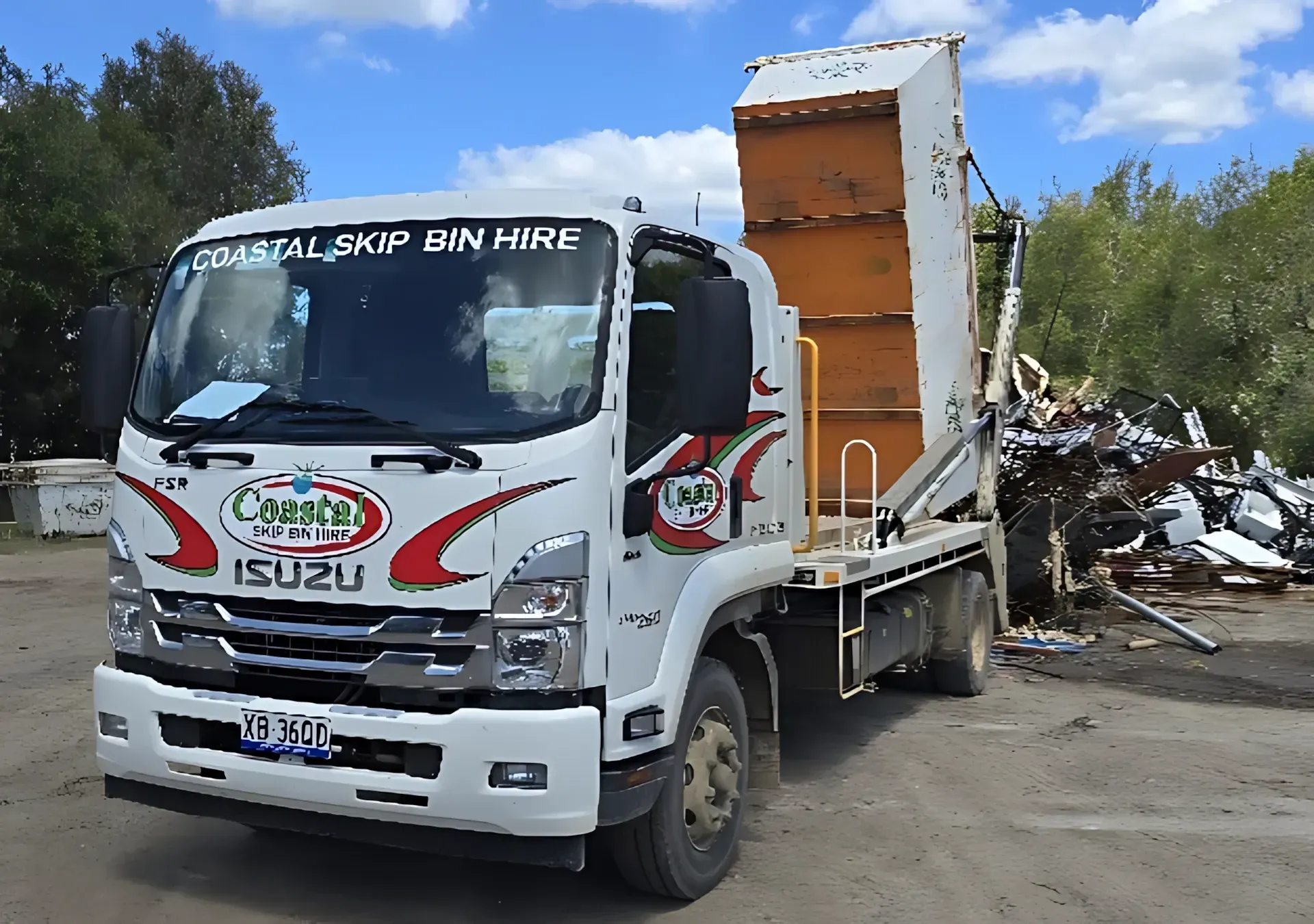 White Isuzu skip bin truck with orange bin, unloading metal scrap. Outdoors on a sunny day — Coastal Skip Bin Hire In Craignish, QLD