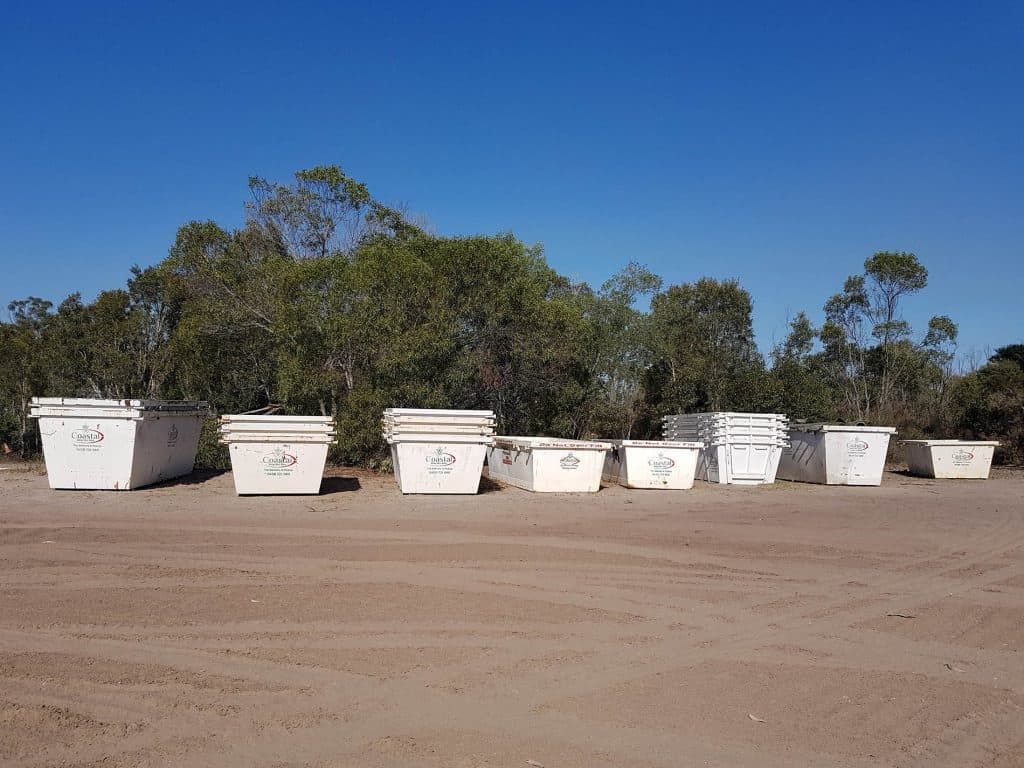 White Dumpsters Lined up On a Dirt Lot in Front of Trees, Under a Clear Blue Sky — Coastal Skip Bin Hire In Craignish, QLD