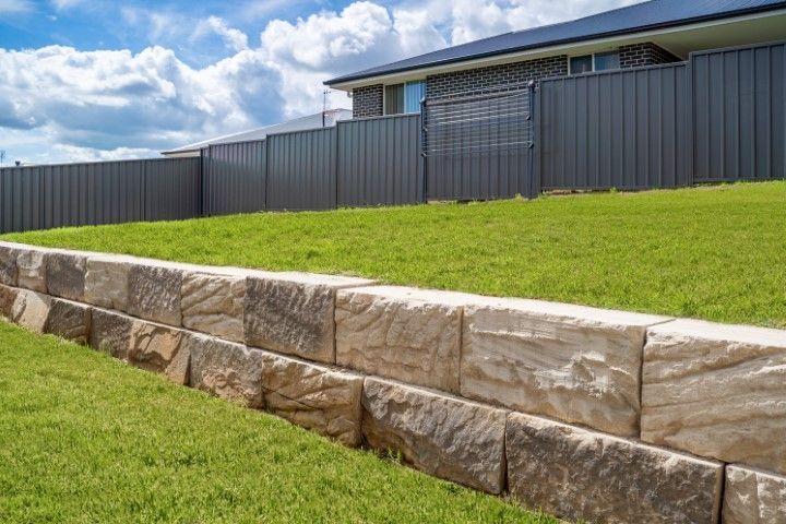 Stone retaining wall beside a grassy slope and dark fence under a cloudy sky
