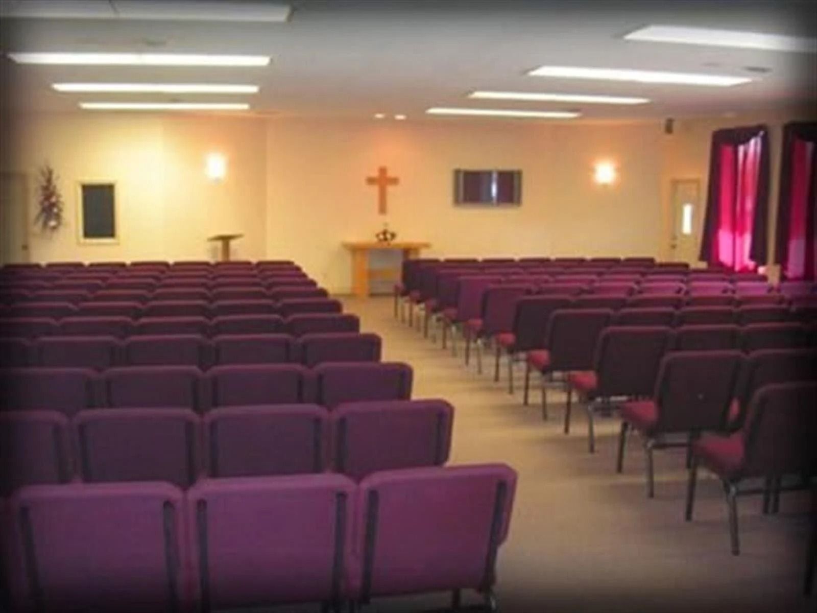 Interior of a church with rows of purple chairs facing an altar with a cross. Red curtains on the right.