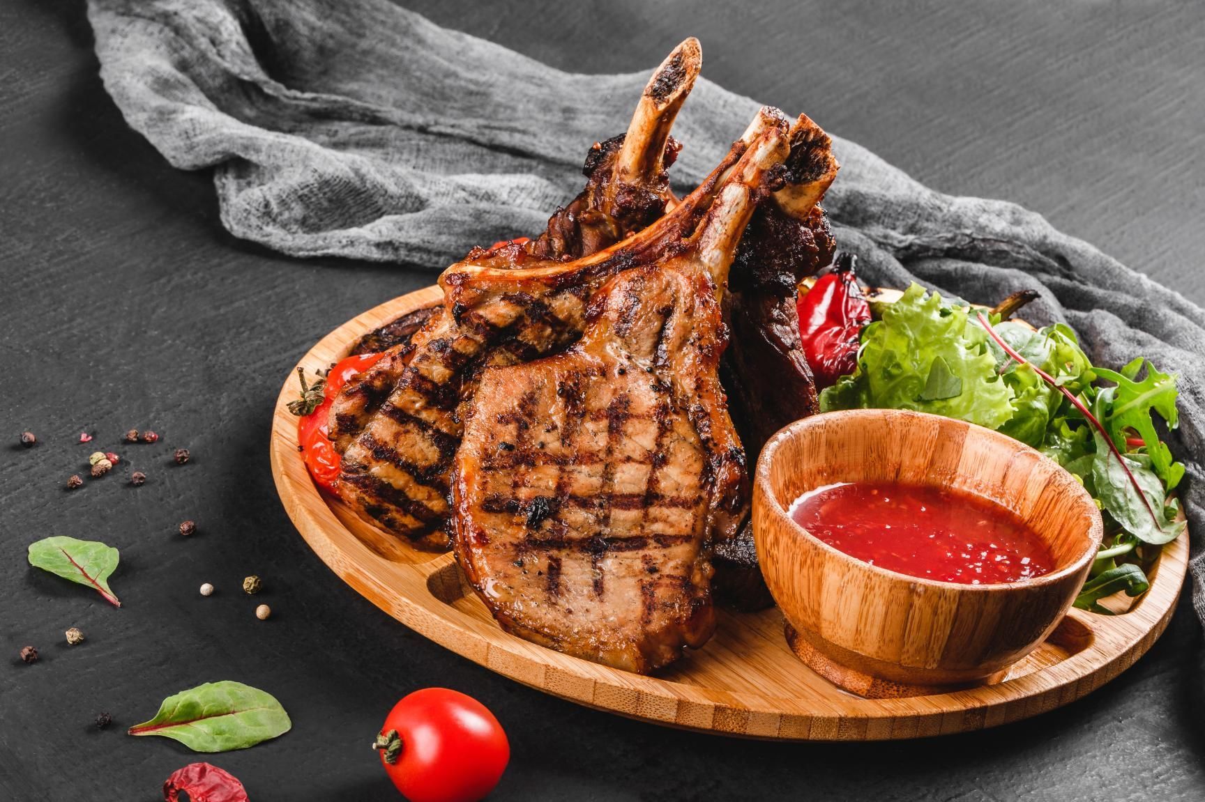 Grilled lamb chops on a wooden board with red sauce, greens, and cherry tomatoes on a dark table