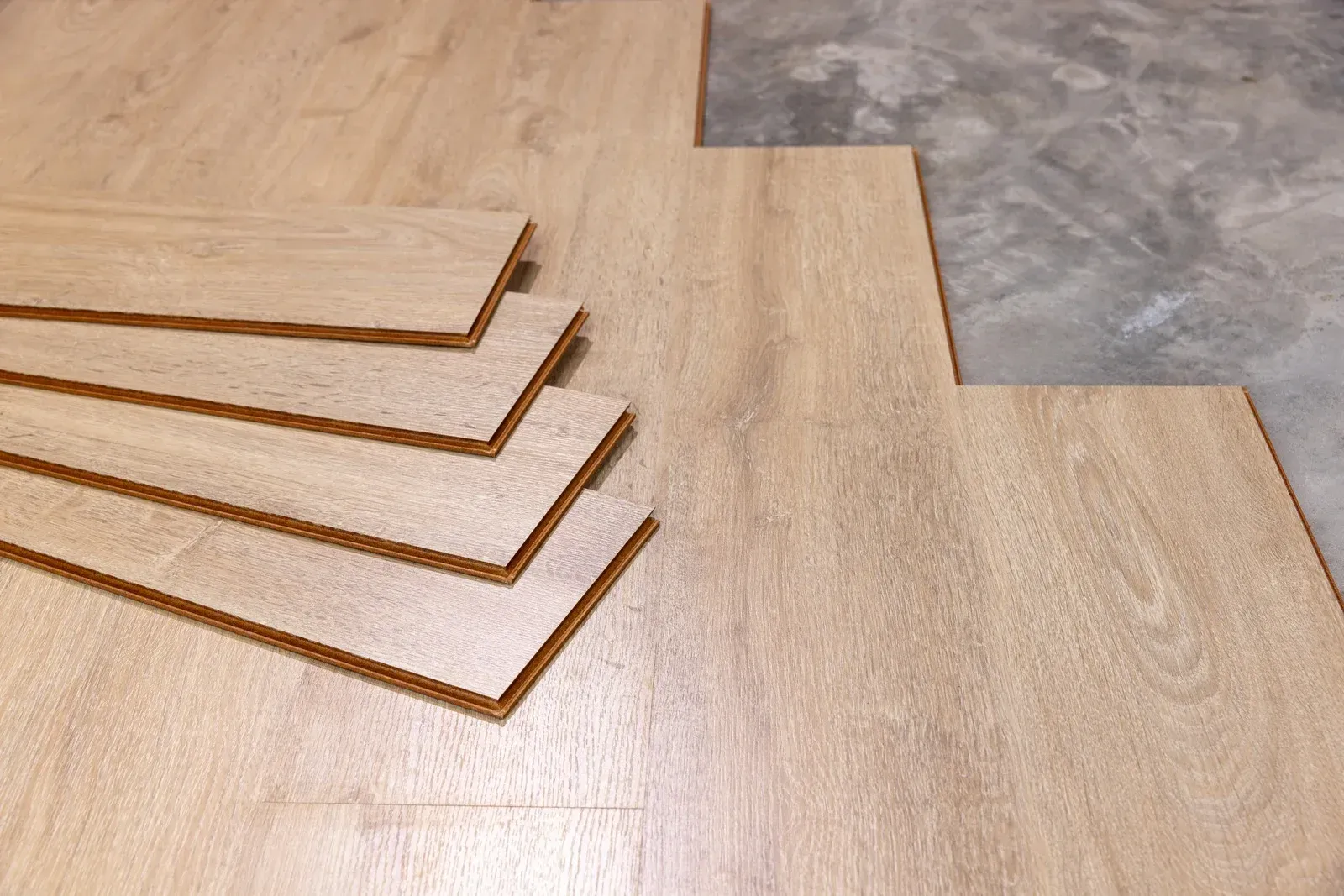Stacked light wood flooring planks on a wood floor beside a gray concrete surface