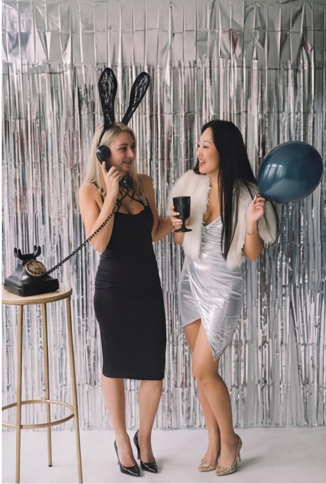 Two women with balloons pose in front of a silver tinsel backdrop. One wears bunny ears, the other a white fur shawl.
