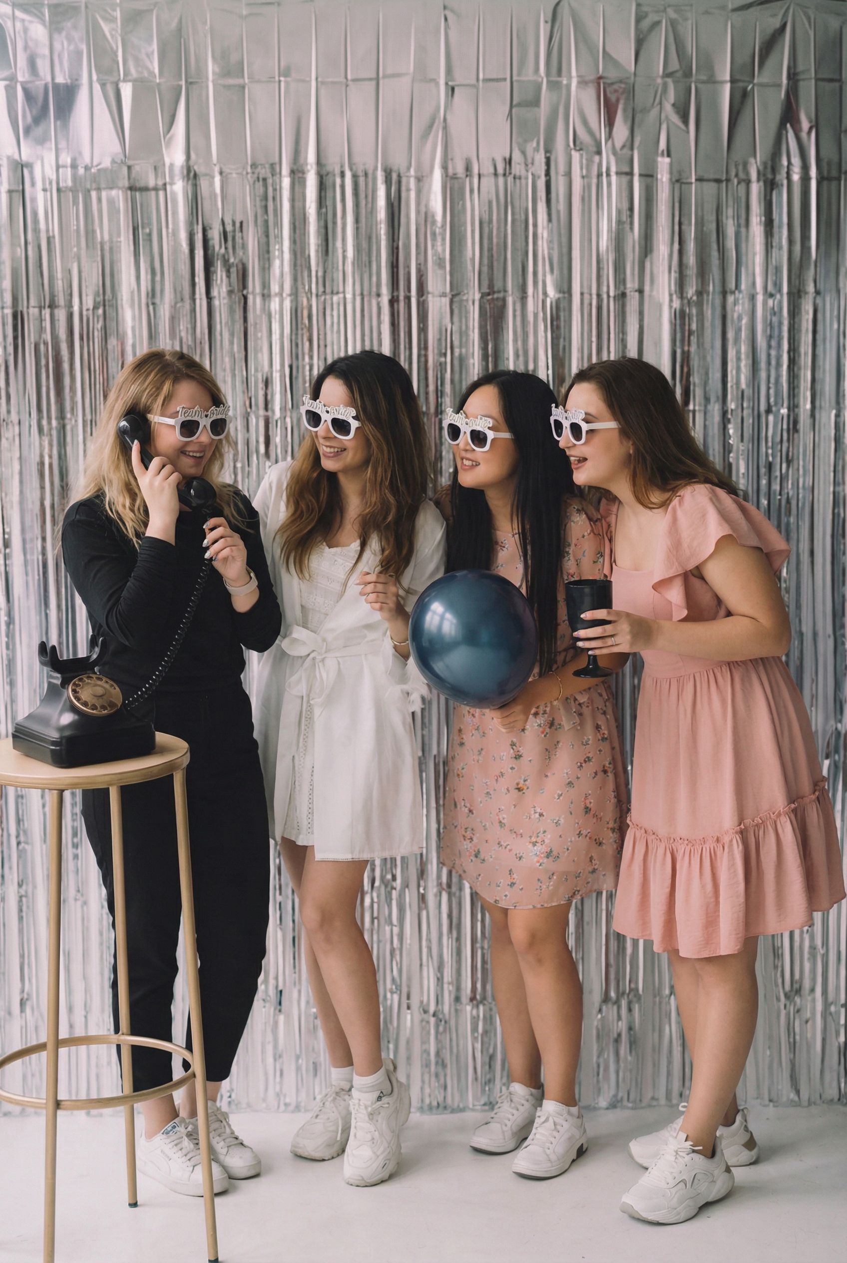 Four women wearing novelty glasses pose with balloons in an ornate room.