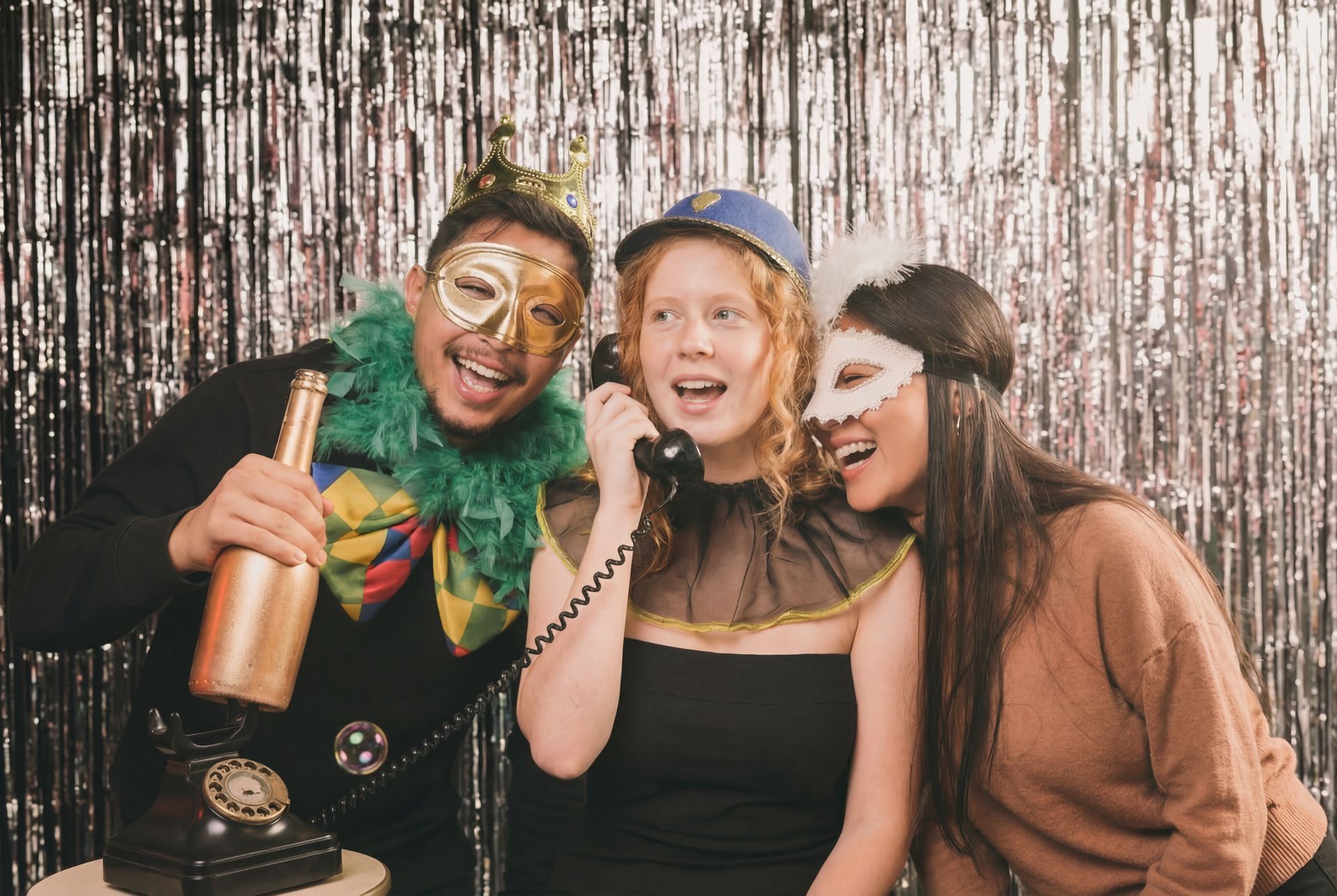 Three people in costume at a party. One holds champagne, another on the phone, and the third laughs. Silver backdrop.