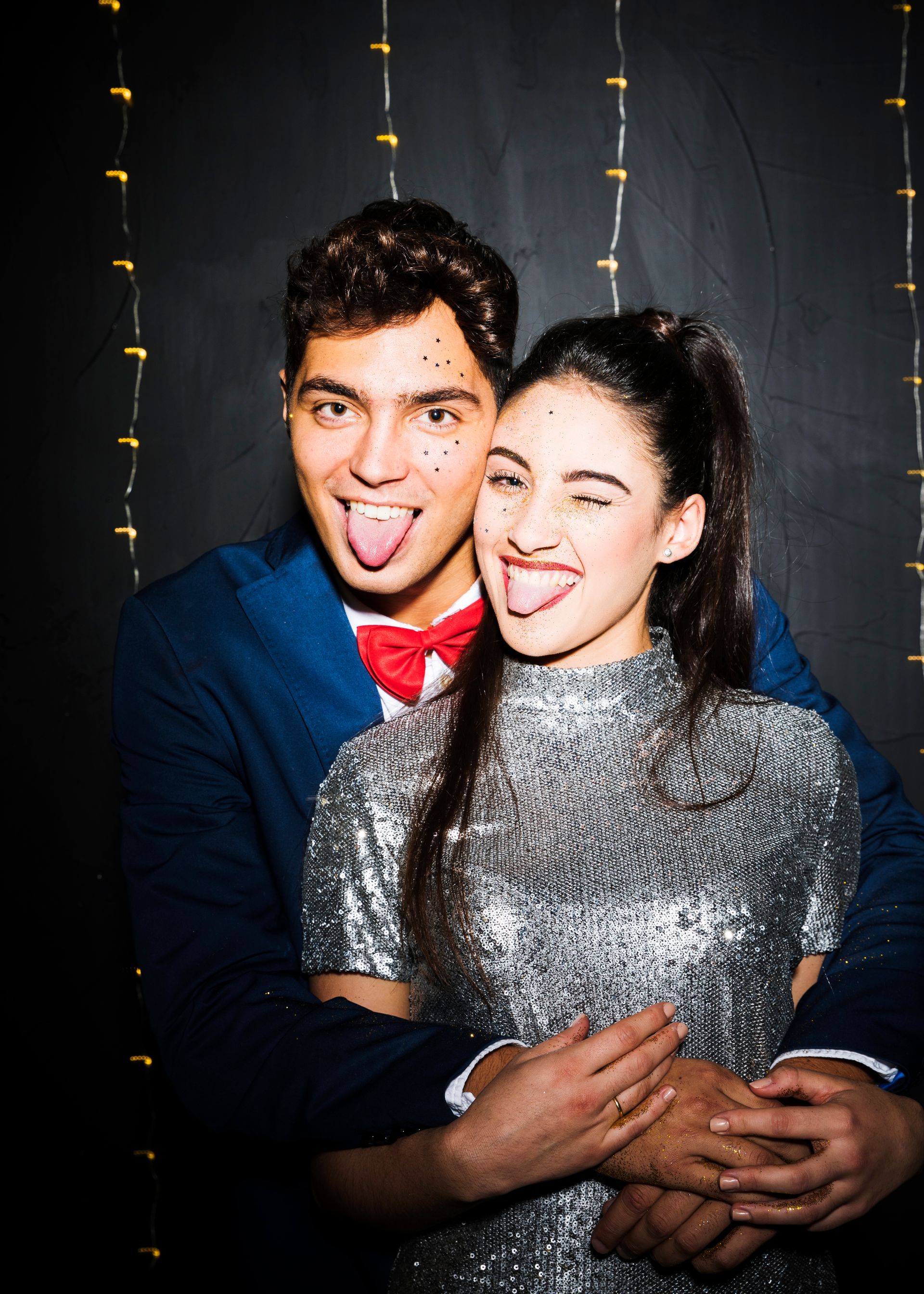 Couple sticking out tongues and winking, posing in front of string lights and a dark backdrop.