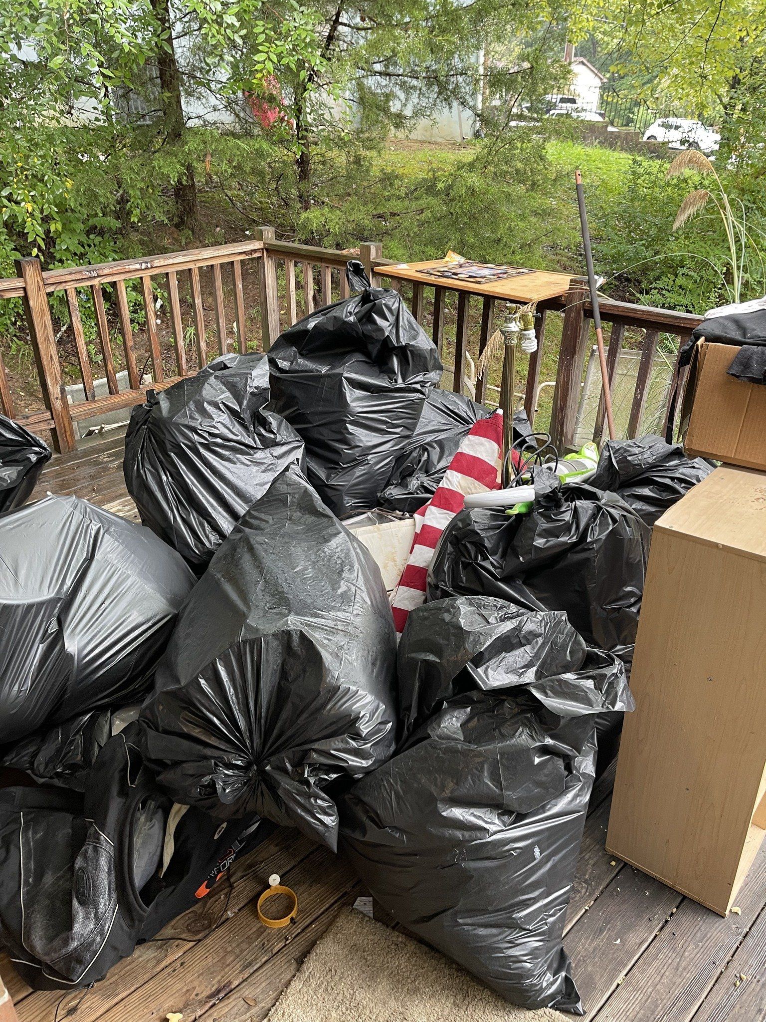 Pile of black garbage bags on a wooden deck with a cardboard box and trees in the background