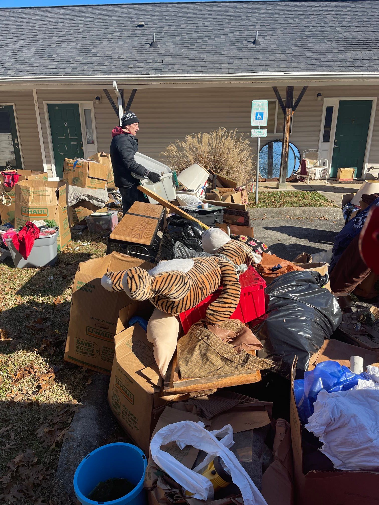 A person in a black coat stacks cardboard boxes and miscellaneous items outside a residential building.