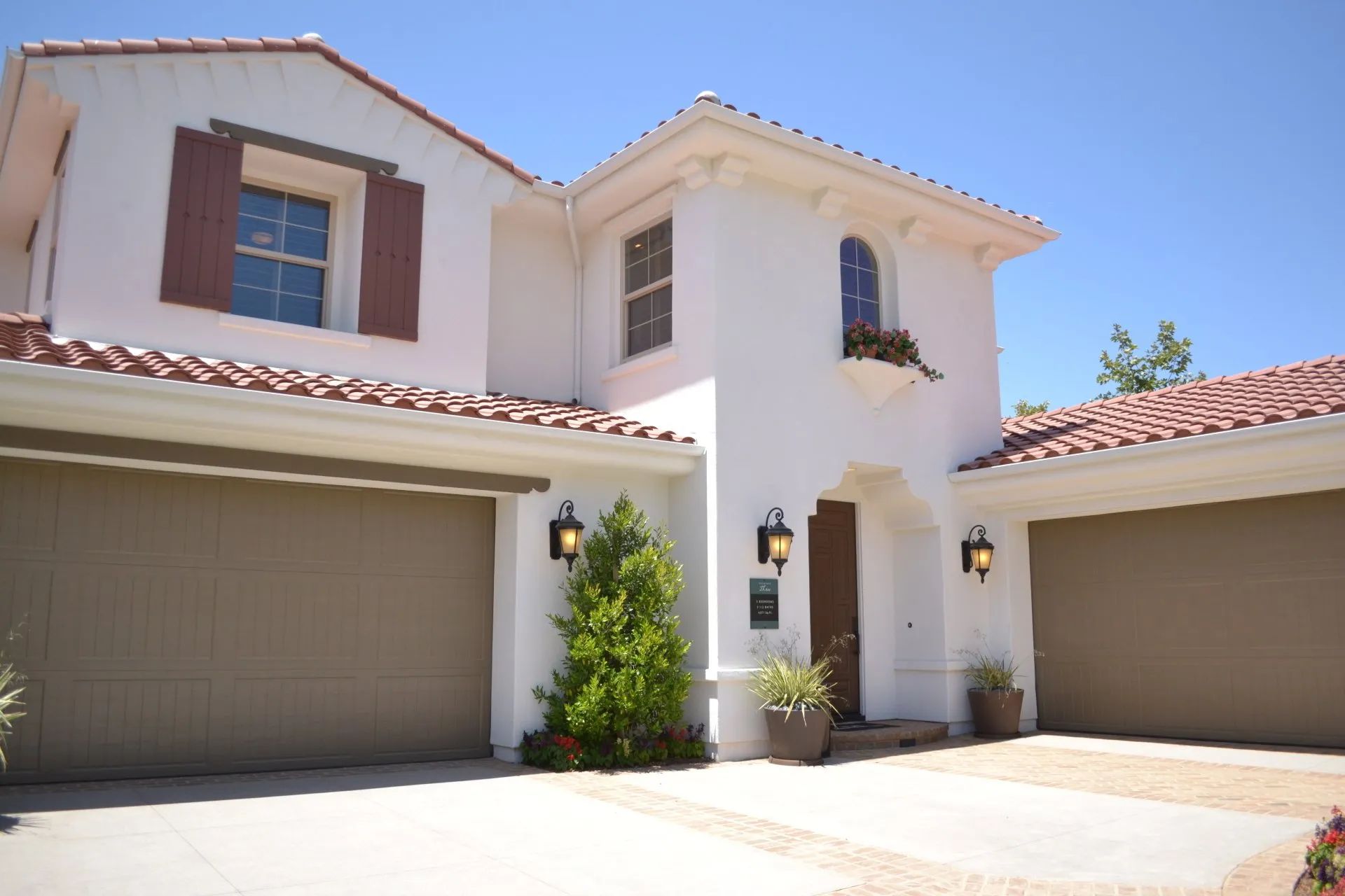 A two-story, white stucco house with a tiled roof, brown garage doors, and a small front entrance with two lanterns.