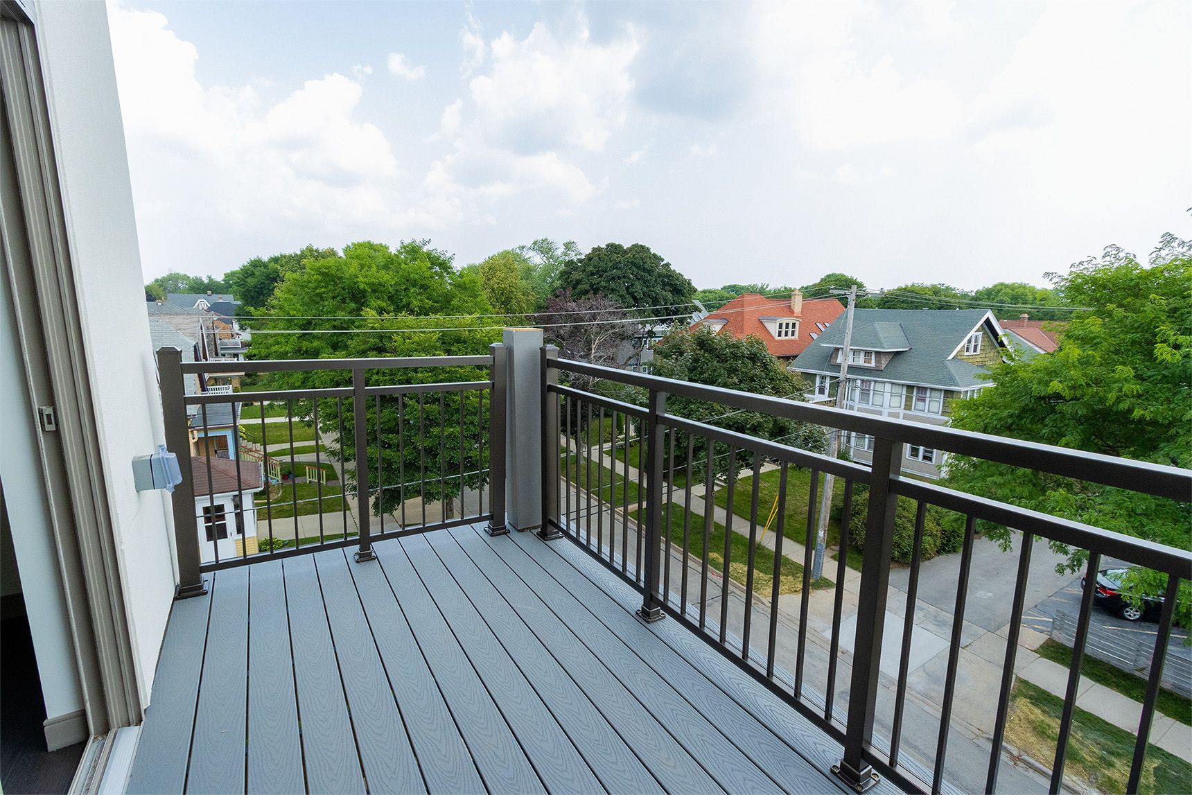 Balcony overlooking a residential neighborhood with green trees and houses, gray deck, metal railing.