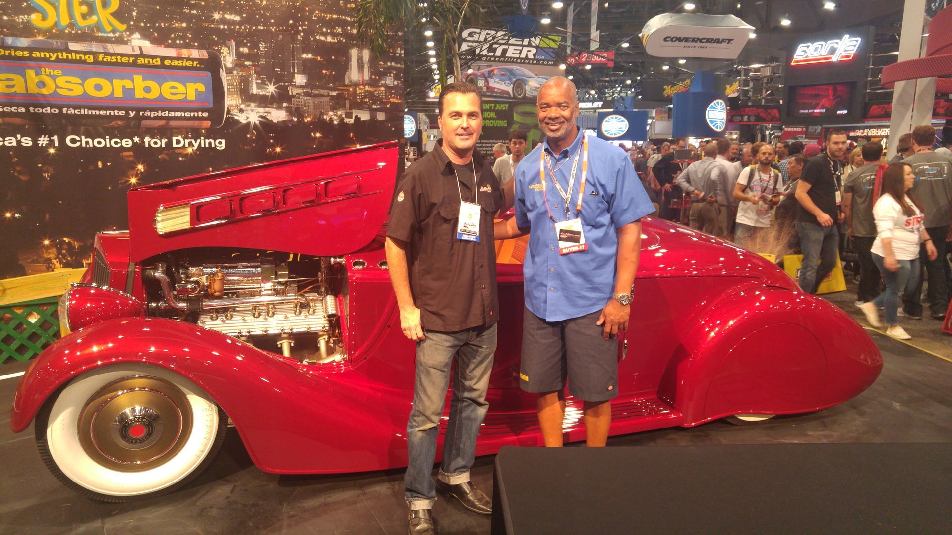 Two men pose with a red custom car at an automotive show; the car has gold wheels and white tires.