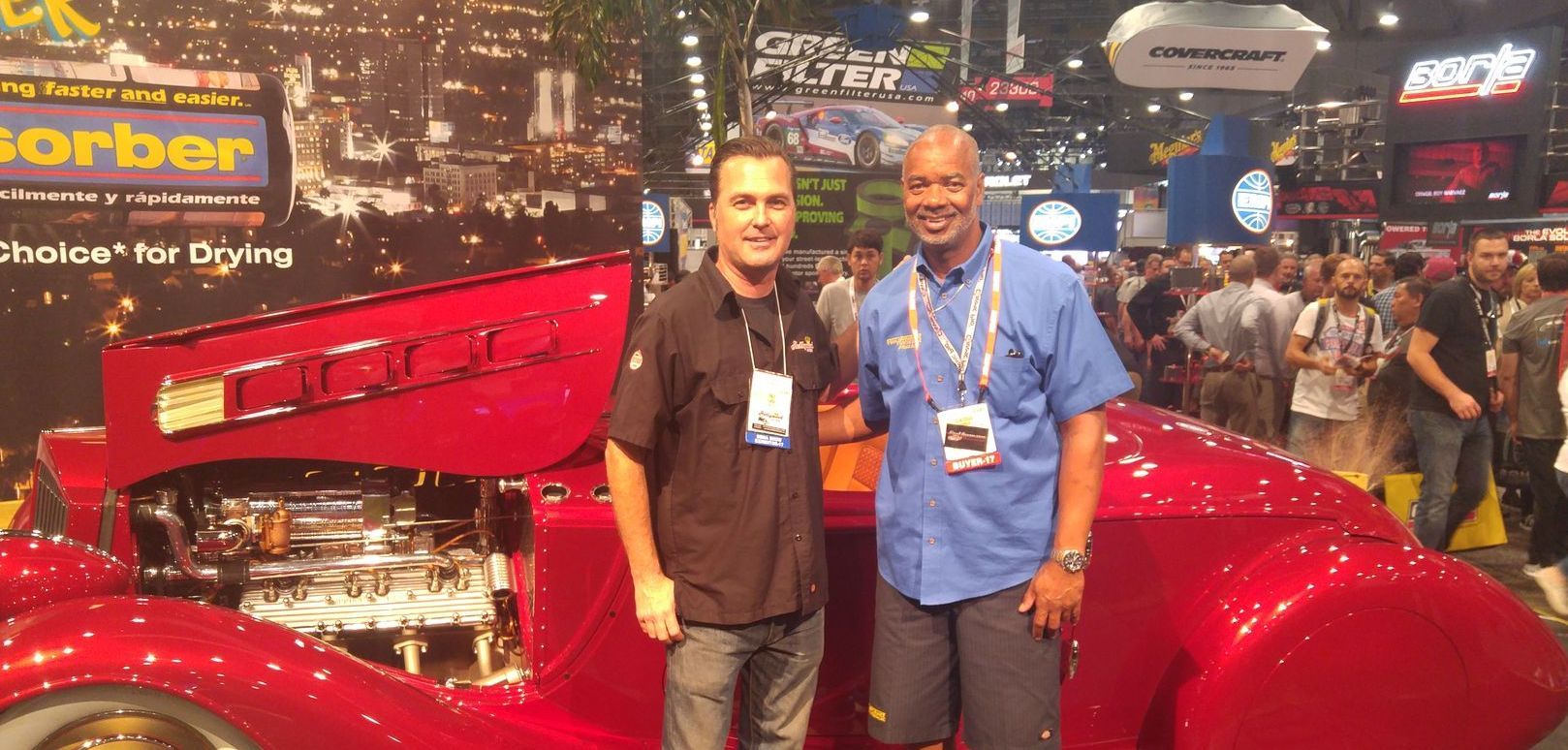 Two men pose by a red vintage car at a car show. They are in front of a cityscape banner, surrounded by a crowd.