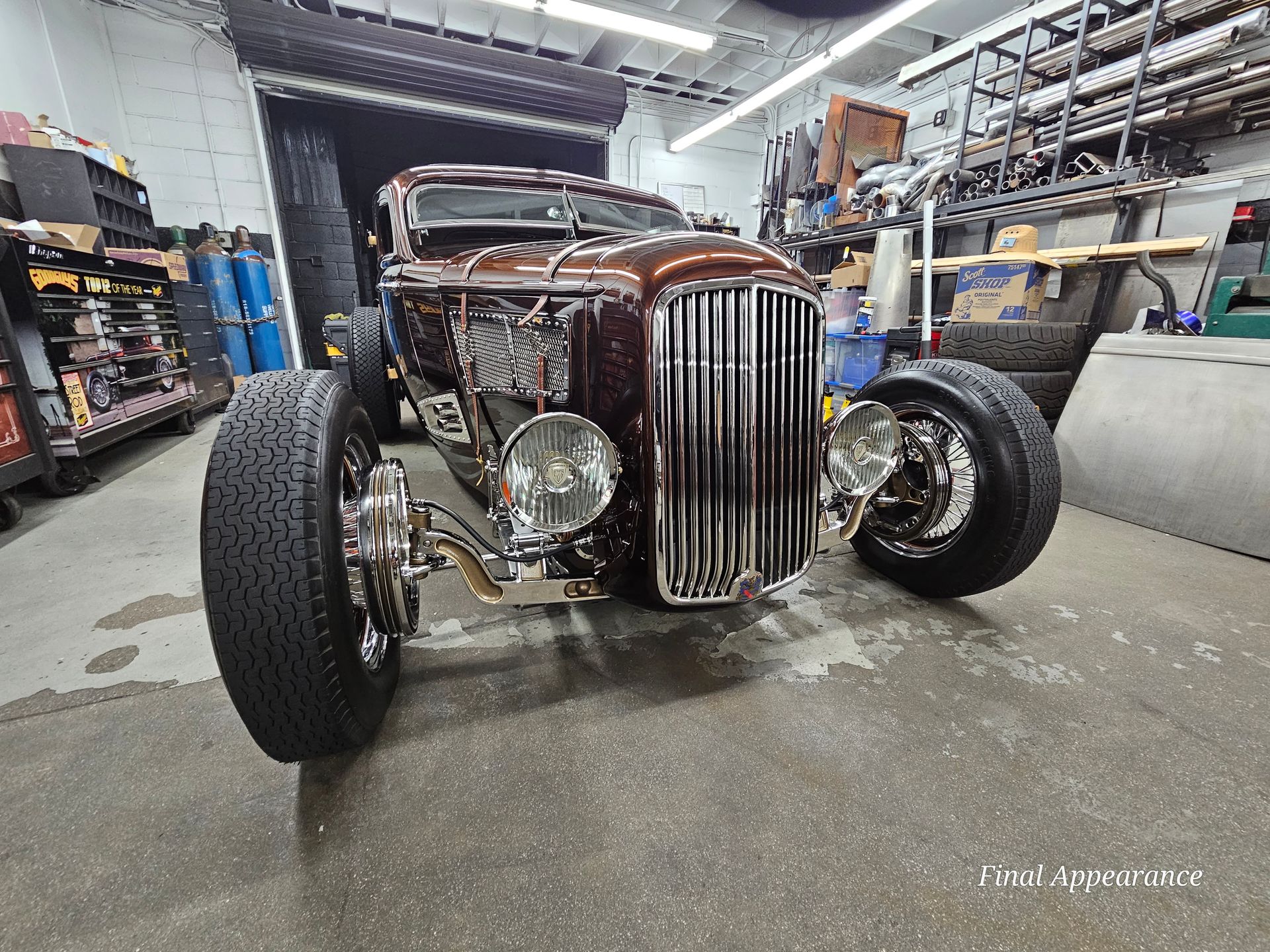 Brown vintage hot rod parked inside a garage. Chrome grill, tires, and headlights.