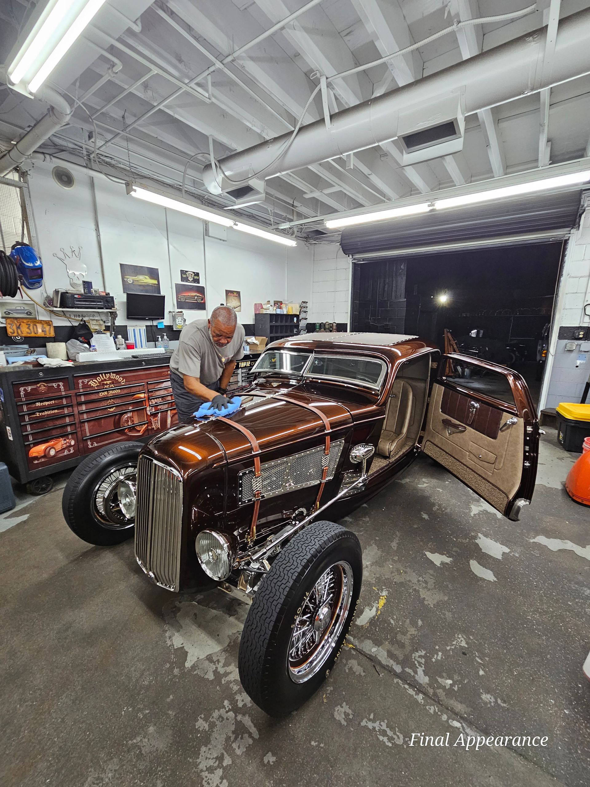 Man polishing a brown, chrome-detailed hot rod in a garage; open doors, tools, and cabinets visible.