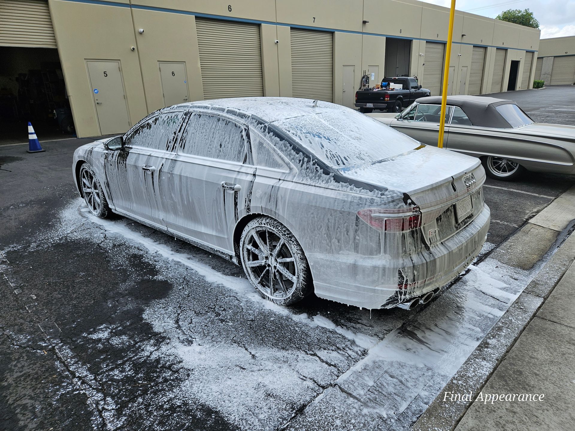 Car covered in white foam at a carwash. Another car parked nearby. Outdoors.