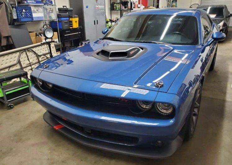 Blue Dodge Challenger with black hood scoop and front splitter, parked in a garage.