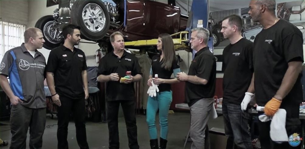 A group of people, including a woman, in a car repair shop. They are talking and standing near a lifted vehicle.