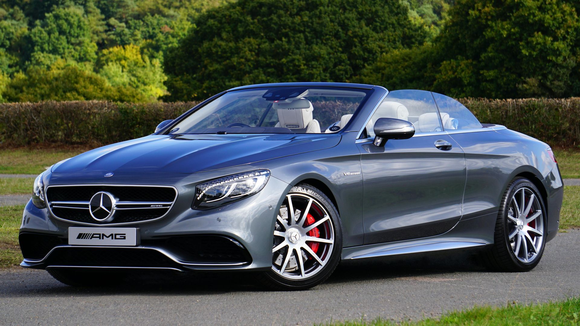 Gray Mercedes-Benz AMG convertible with red brake calipers parked on a paved road, trees in the background.