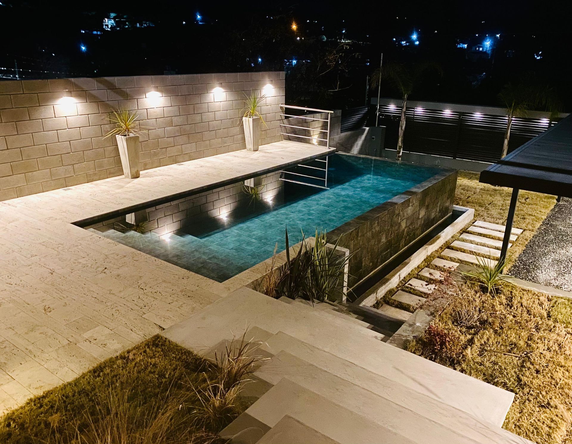 Vista nocturna de una piscina rectangular con luces, un muro de piedra y escalones que conducen al área de la piscina.