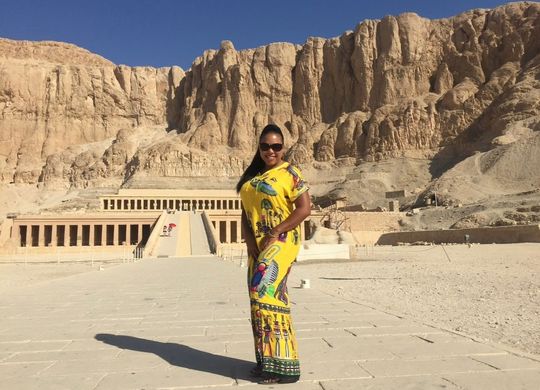 Woman in yellow dress poses at the Temple of Hatshepsut in Egypt, with beige mountains in the background.