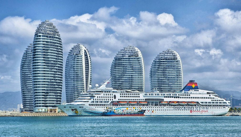 Cruise ship docked in front of Sanya Atlantis resort in China, with unique spiral-shaped buildings against a cloudy sky.