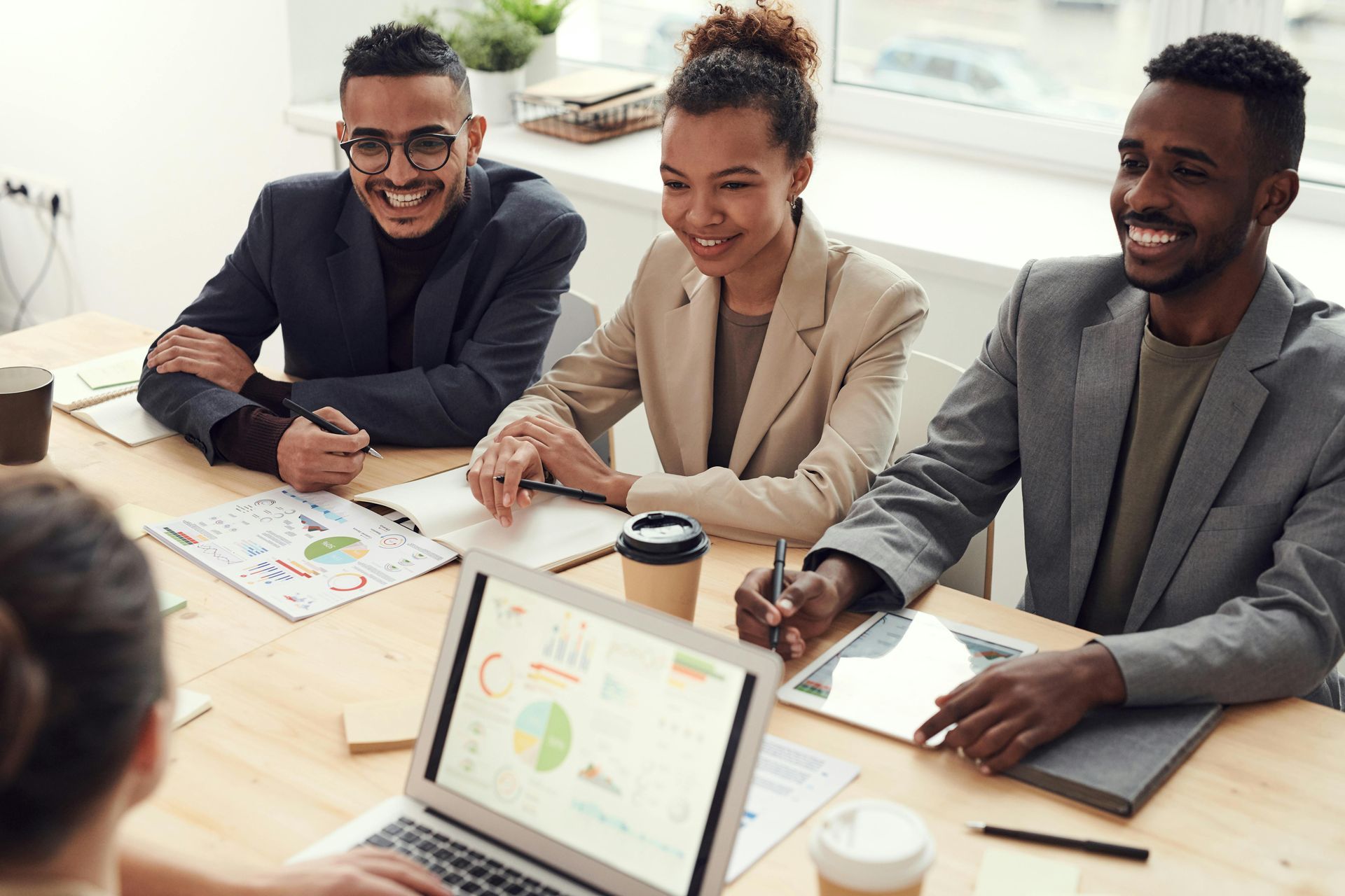Four colleagues in a meeting, smiling around a laptop with charts. Natural light, beige table.