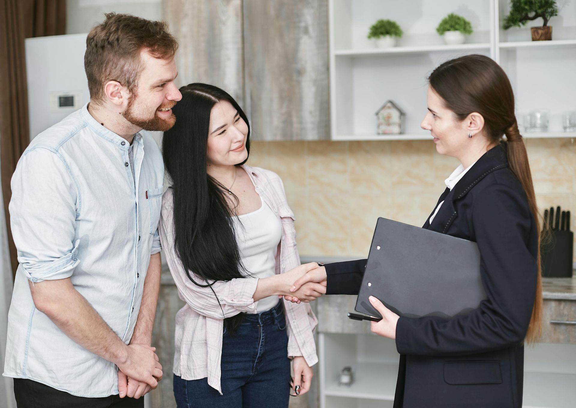 Couple shaking hands with realtor in a modern kitchen; smiling faces, document folder.