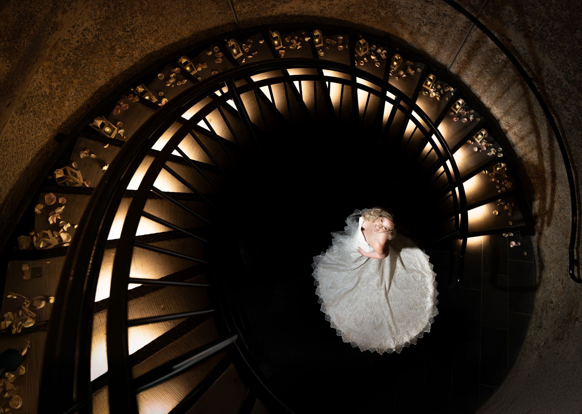 Bride in white dress on spiral staircase, lit from above, looking down.