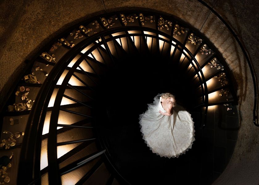 Bride in white dress on spiral staircase, lit from above, looking down.