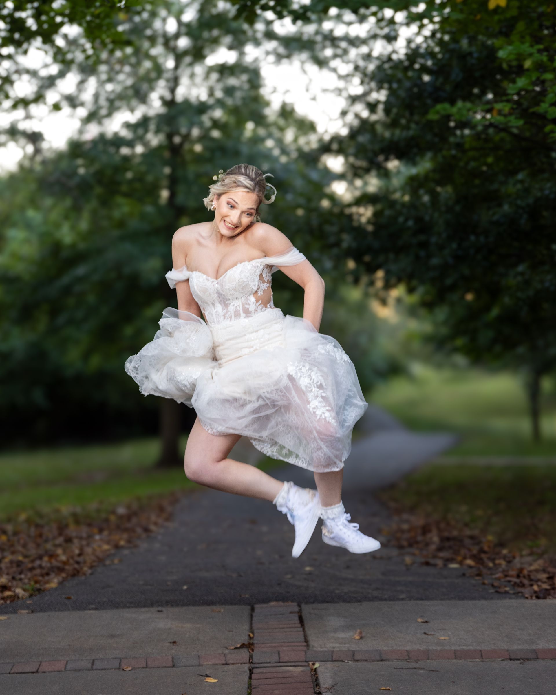Woman in a white dress and sneakers jumping on a path in a park, smiling.
