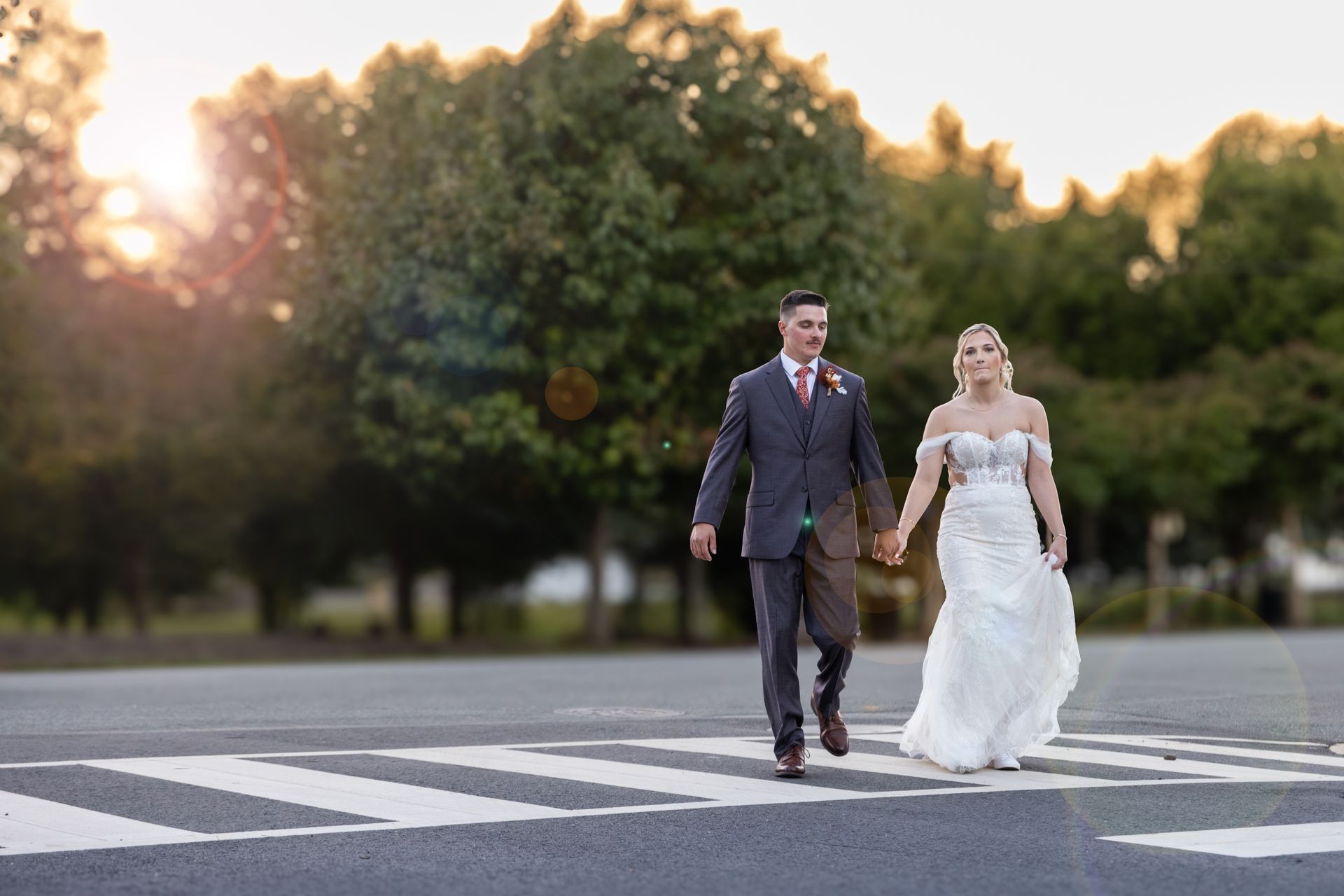 Bride and groom hold hands, crossing a crosswalk. Sun sets behind them, trees in the background.