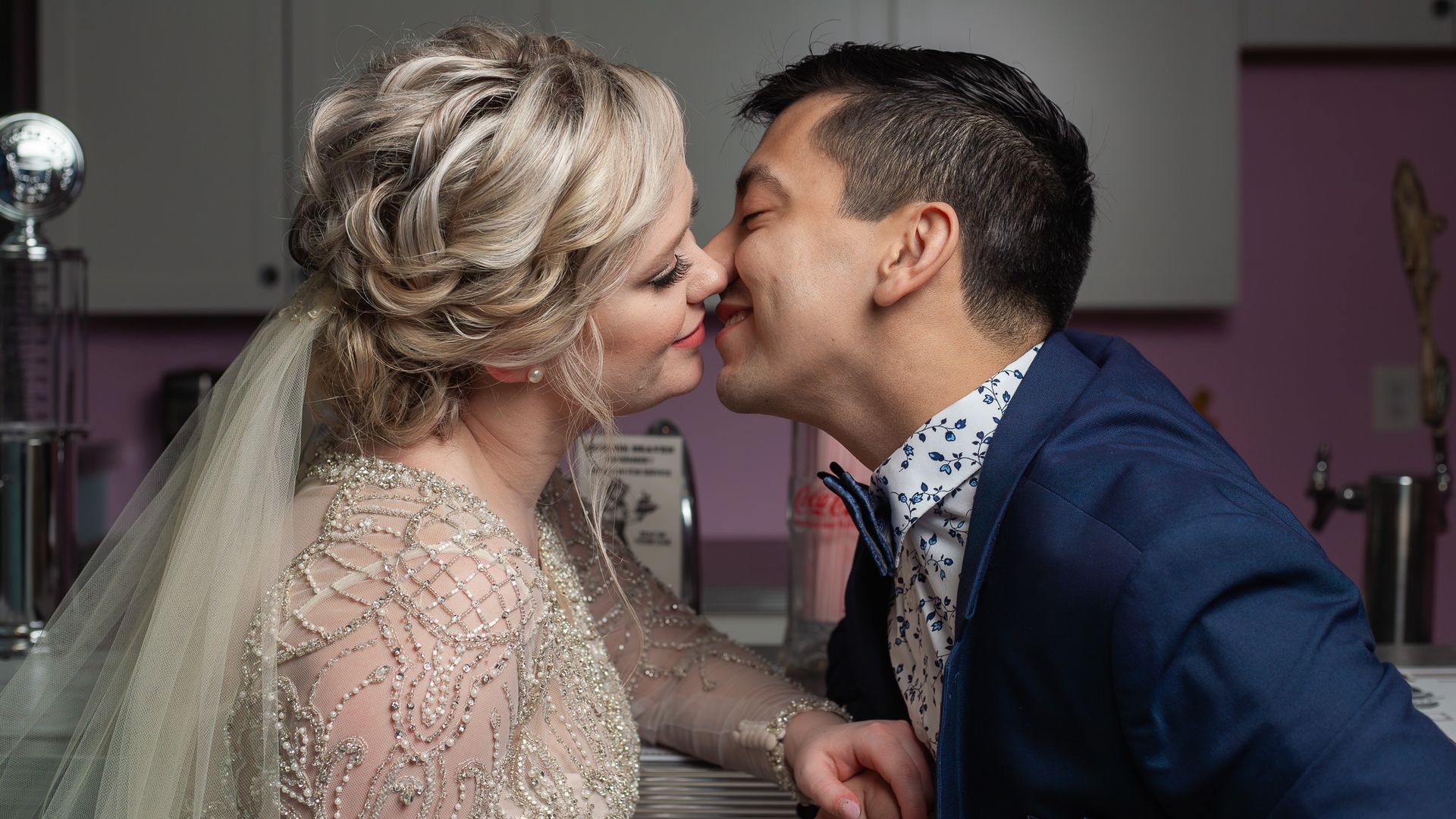 Bride and groom kissing, leaning over a counter. Bride wears a veil and gown, groom wears a blue suit.