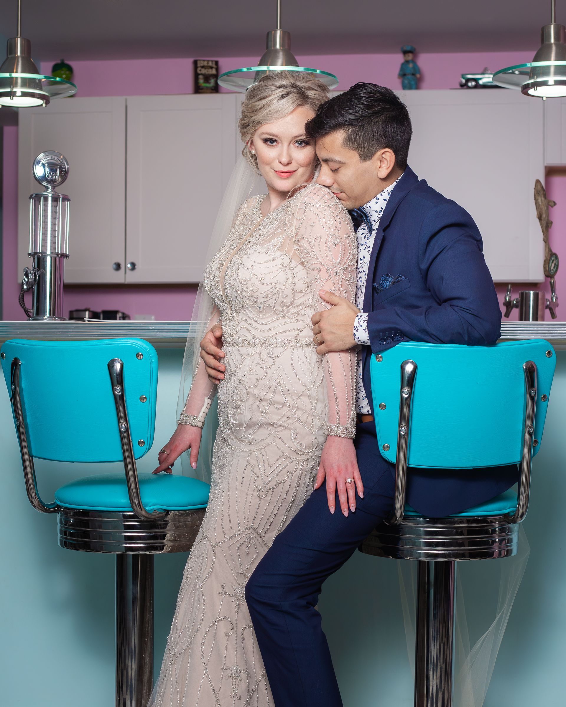 Bride and groom embracing by a retro diner counter. Bride in gown, groom in suit. Turquoise barstools.
