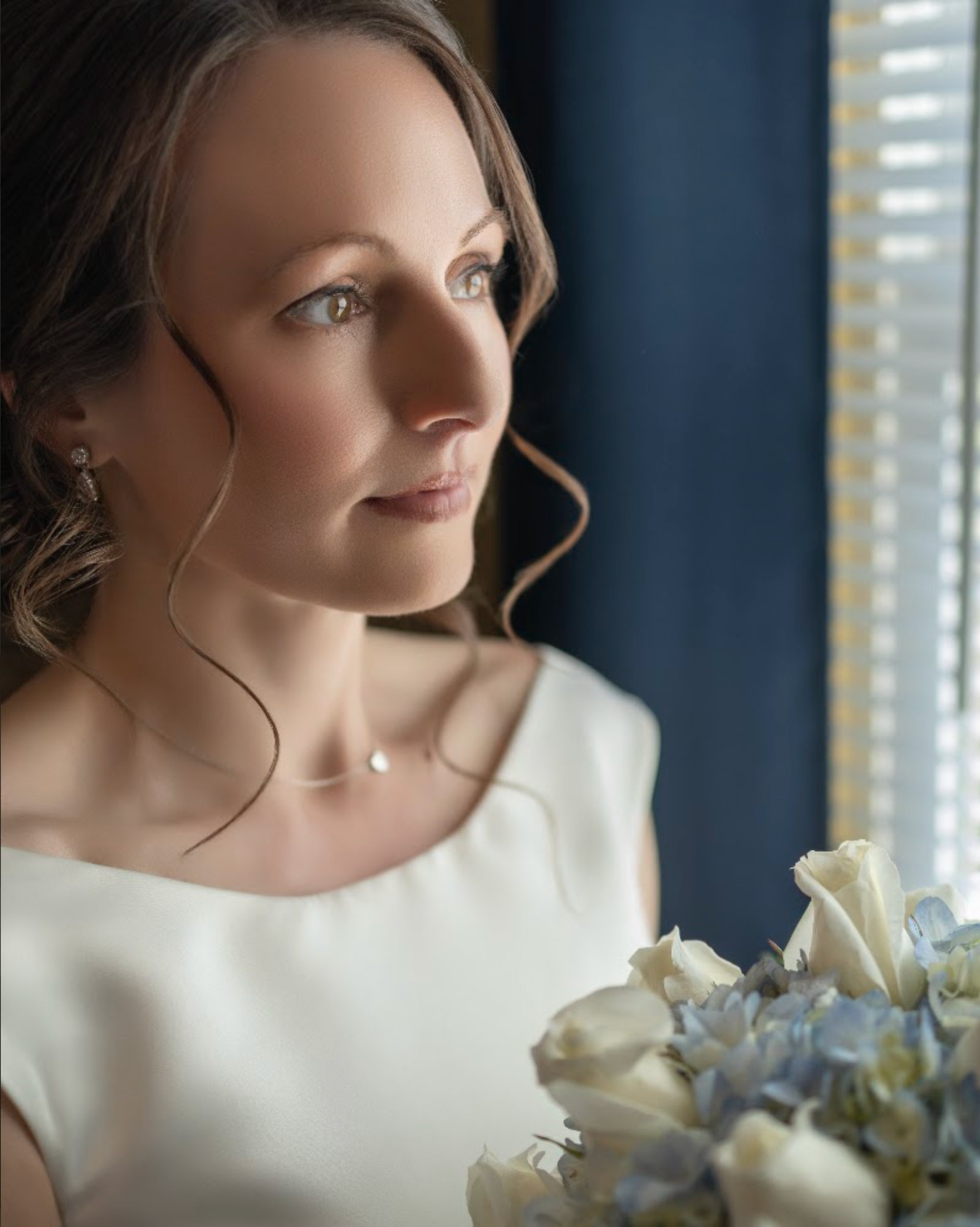 Woman in a white dress holds flowers, looking out a window.