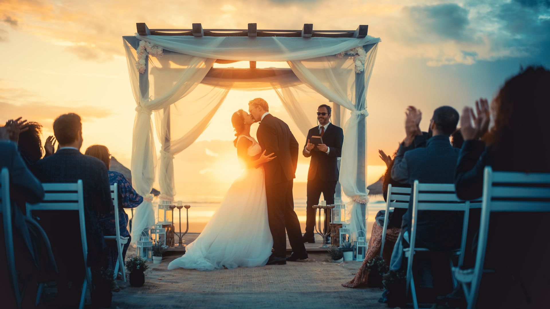 Couple kissing at sunset wedding ceremony, guests clapping, ocean setting.