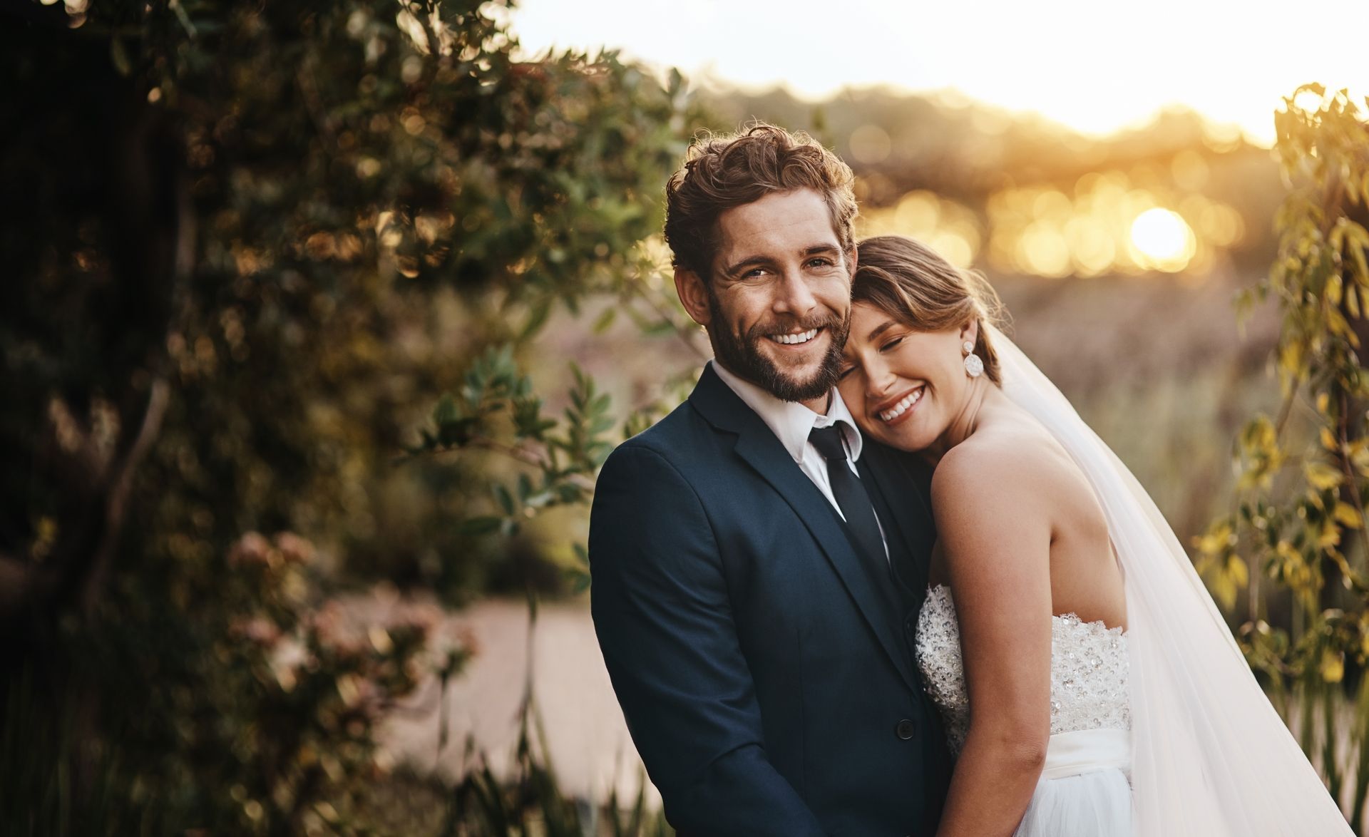 Smiling couple on their wedding day, embracing outside; sunlight.