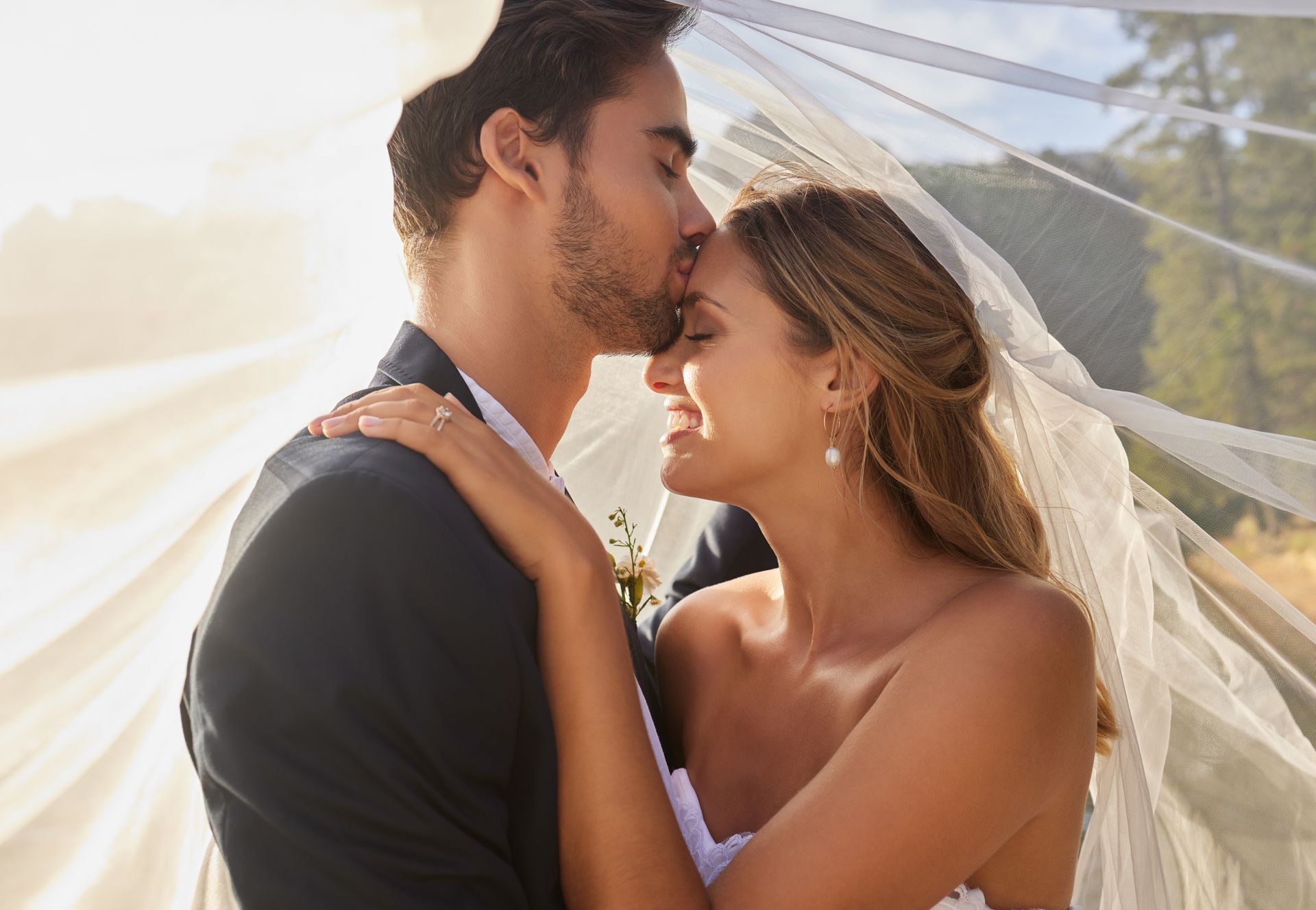 Groom kissing bride's forehead, both smiling under a veil in sunny outdoor setting.