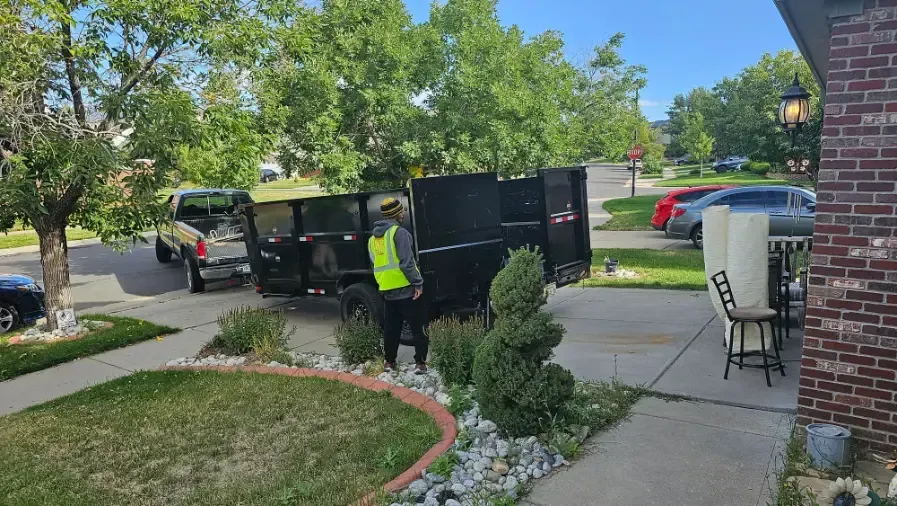 Man in safety vest near a black dumpster being loaded by a truck in a suburban driveway.