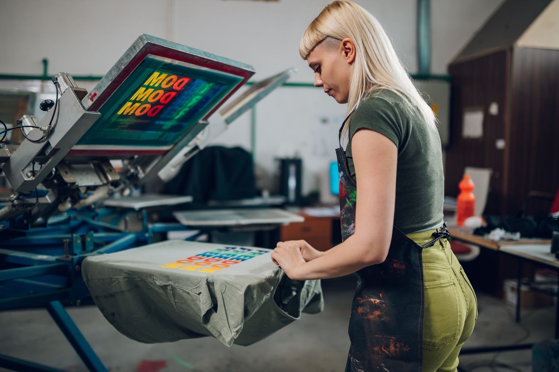 Woman using custom screen printing services to print colorful design on a t-shirt in workshop.