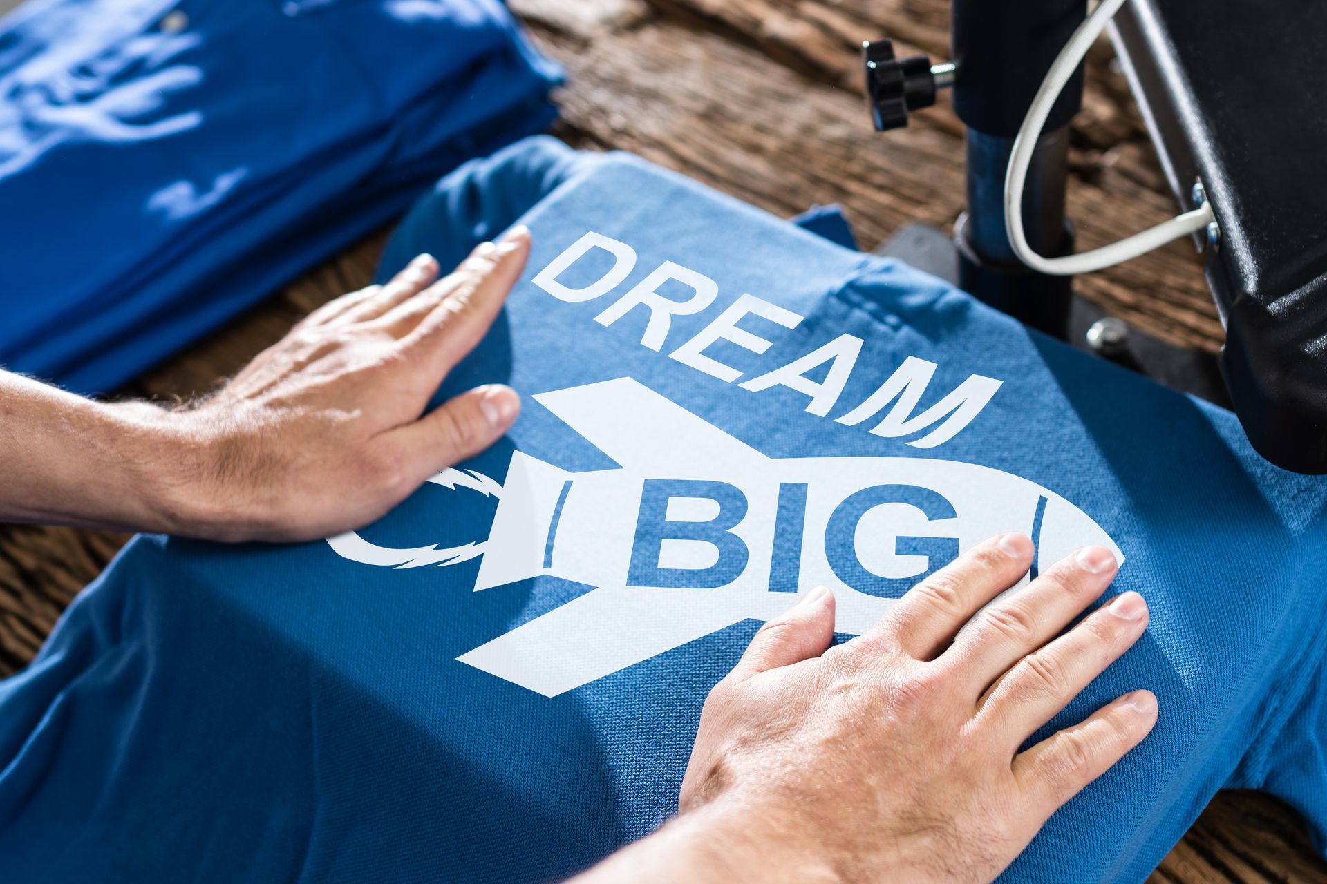 Person pressing blue t-shirt with 'Dream Big' rocket design using heat press machine.