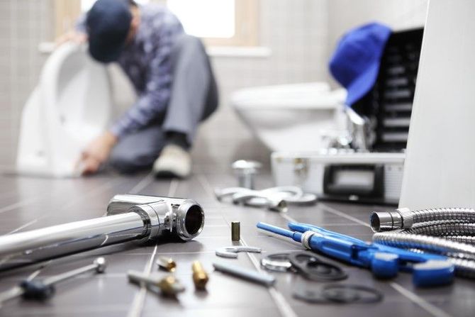 A plumber works on a toilet in a bathroom, with tools and plumbing components scattered on the tiled floor in the foreground.